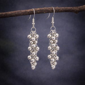 May include: A pair of silver-toned dangle earrings featuring a cluster of small, round, metallic beads suspended from a thin wire hook. The beads are arranged in a cascading, multi-layered formation that tapers towards the bottom of each earring.