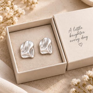 May include: A pair of silver-toned earrings with an irregular, organic, wavy surface texture. The earrings are presented inside a beige cardboard gift box with the text A little brighter every day and a small heart symbol printed on the lid.
