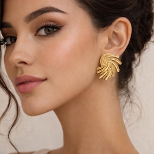 Puede incluir: Primer plano de una mujer con pendientes dorados en forma de espiral. Los pendientes tienen un diseño texturizado y acanalado. La mujer tiene el pelo oscuro y está maquillada. Los pendientes son un rasgo destacado.