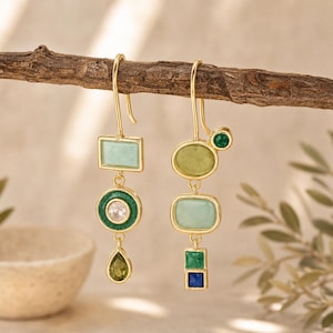 May include: A pair of gold-tone drop earrings featuring mismatched geometric shapes. The left earring has a light green rectangle, a dark green circle with a clear centre stone, and a teardrop gem. The right earring has an oval, a light green rectangle, and two small square stones.