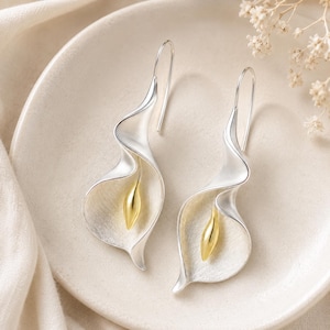 May include: A pair of drop earrings shaped like calla lilies, featuring a matte silver finish on the petals and a polished gold-tone centre spadix. Each earring hangs from a simple silver hook, showcasing a curved, organic floral design.