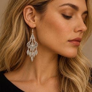 May include: A silver-toned chandelier earring featuring an intricate filigree design with multiple teardrop-shaped dangles. The metalwork displays a series of concentric oval patterns and small spherical accents, suspended from a simple hook wire.