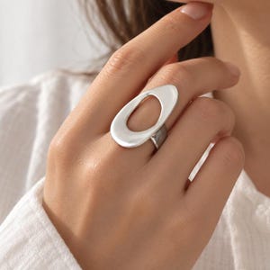 May include: A silver-toned ring featuring a large, smooth, asymmetrical oval-shaped face with a hollow center. The band is polished and connects to the base of the wide, organic-shaped top piece, creating a bold and prominent statement piece.