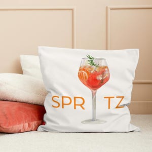 Könnte beinhalten: Ein weißes Kissen mit einer orangefarbenen und roten Illustration eines Glases Spritz mit einem Zweig Rosmarin. Der Text "SPRITZ" ist in Orange unter dem Glas gedruckt.