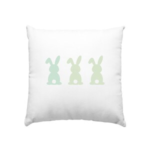 Könnte beinhalten: Weißes quadratisches Kissen mit drei Hasen-Silhouetten in einem hellgrünen und blauen Farbverlauf. Die Hasen sind horizontal über die Mitte des Kissens angeordnet und schaffen ein einfaches, aber charmantes Design.
