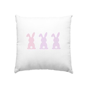 Könnte beinhalten: Weißes quadratisches Kissen mit drei Hasensilhouetten in Pastellrosa und Lila. Die Hasen sind horizontal über das Kissen angeordnet, von hinten betrachtet. Das Kissen hat ein weiches, flauschiges Aussehen.