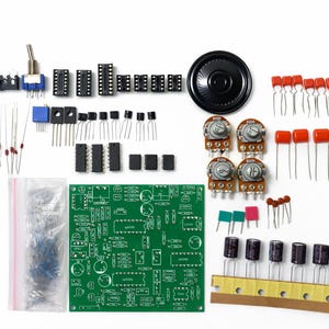 Może przedstawiać: Zestaw dla hobbystów elektroniki zawierający zieloną płytkę drukowaną, czarny głośnik, różne układy scalone, tranzystory, potencjometry, kondensatory, rezystory, przełącznik dźwigniowy oraz plastikową torebkę z drobnymi elementami elektronicznymi.
