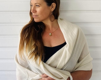 May include: A woman wearing a white shawl, a black tank top, and a brown necklace with a small pendant. She is also wearing a brown woven oval-shaped earring.