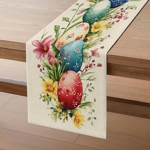 May include: A rectangular Easter table runner featuring watercolor-style painted Easter eggs in shades of red, blue, and yellow, surrounded by flowers and greenery. The runner is made of a light-colored fabric.