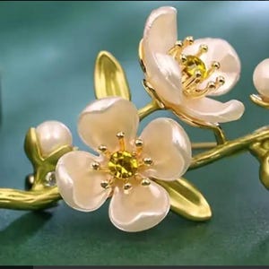 May include: A gold-tone brooch featuring a delicate branch with two white blossoms and pearl accents. The blossoms have yellow centers and a pearlescent finish.