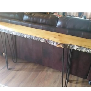 May include: A long, natural wood console table with a live edge top and black hairpin legs. The table is in front of a brown leather sofa.