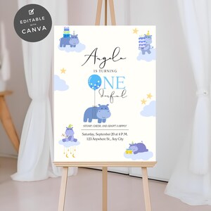 Könnte beinhalten: Ein weißes Schild auf einer Staffelei mit Geburtstagsthema. Das Schild zeigt eine blaue Nilpferd-Illustration, den Text „Angela wird eins“ und Veranstaltungsdetails. Das Design umfasst Wolken, Sterne und einen Ballon.
