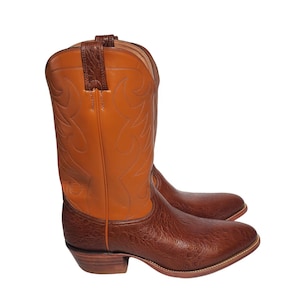 Op de afbeelding: Een paar tweekleurige leren cowboylaarzen met een donkerbruine, getextureerde voet met een generfde afwerking en een lichtere, tan-kleurige schacht. De schacht is versierd met decoratieve ton-sur-ton stiksels en heeft treklussen aan de bovenkant.