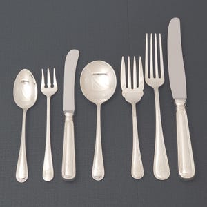 Könnte beinhalten: Eine Sammlung silberfarbener Besteckteile, darunter Löffel, Gabeln und Messer, auf einer dunkelgrauen Oberfläche angeordnet. Die Utensilien haben ein klassisches Design mit abgerundeten Griffen und dekorativen Details. Das Set beinhaltet einen Servierlöffel und ein Buttermesser.
