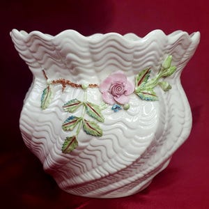 May include: A white ceramic planter with a wavy design and a raised floral motif. The planter features a pink rose with green leaves.