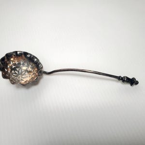 May include: A silver-toned metal sifter spoon featuring a scalloped, bowl-shaped head with intricate cut-out floral patterns. The handle is slender and curved, terminating in a decorative, textured finial with a dark, tarnished patina finish.