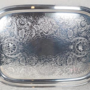 May include: A silver serving tray with ornate embossed designs and two handles. The tray is oval shaped and has a shiny finish.