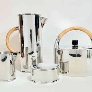 May include: A collection of polished silver serving items. It includes a tall jug with a woven handle, a teapot with a matching handle, a small creamer, and a sugar bowl. The teapot and jug have a sleek, modern design.