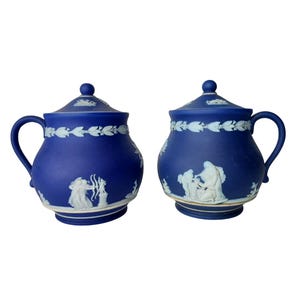 May include: Two cobalt blue ceramic lidded vessels featuring white relief ornamentation. Each piece displays a handle, a lid with a knob, and a decorative band of leaves around the top, with classical figures depicted in white on the main body.