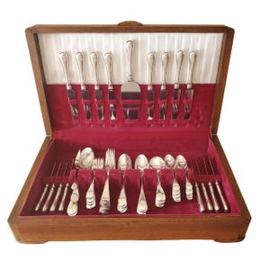 Peut inclure: Un coffret en bois pour couverts avec un intérieur en velours rouge, contenant un ensemble complet de couverts de couleur argentée. L'ensemble comprend des couteaux, des fourchettes, des cuillères et un serveur à gâteau, tous soigneusement rangés.