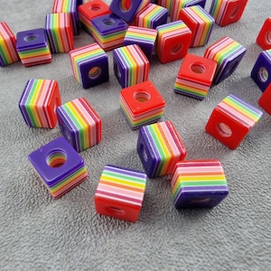 Könnte beinhalten: Eine Sammlung farbenfroher, würfelförmiger Perlen. Die Perlen sind hauptsächlich rot und lila, mit Regenbogenstreifen an den Seiten. Jede Perle hat ein Loch in der Mitte, das zum Auffädeln geeignet ist. Diese Perlen sind ideal für die Schmuckherstellung.