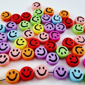 Könnte beinhalten: Eine Sammlung bunter Smiley-Gesichtsperlen in verschiedenen Farben, darunter Orange, Rot, Rosa, Lila, Gelb, Grün und Blau. Jede Perle hat ein schwarzes Smiley-Gesicht-Design. Geeignet für Schmuckherstellung und Bastelarbeiten.