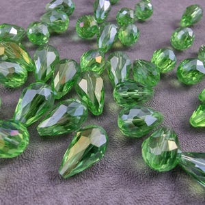 Könnte beinhalten: Eine Sammlung grüner Glasperlen in Tropfenform. Die Perlen sind facettiert und haben einen leichten irisierenden Schimmer.