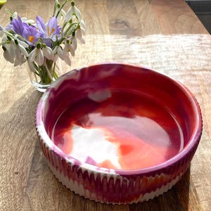 Op de afbeelding: Een ronde, decoratieve kom met een levendig, rood, roze en wit gemarmerd interieur. De kom heeft een getextureerde, geribbelde buitenkant met een wit en roze patroon. Een kleine glazen vaas met paarse en witte bloemen staat in de buurt.