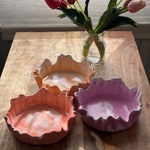 Könnte beinhalten: Drei runde, flache Schalen mit welligen, gewellten Rändern. Eine Schale hat ein marmoriertes pfirsich-weißes Muster, eine andere ein orange-weißes Design und die dritte eine marmorierte rosa-weiße Oberfläche mit einer glatten Textur.