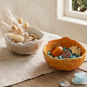 Könnte beinhalten: Zwei dekorative Keramikschalen. Eine ist beige und mit Kieselsteinen und getrockneten Pflanzen gefüllt. Die andere ist orange und enthält türkisfarbene Steine, eine Spirale und ein goldenes Blatt. Zusätzliche dekorative Elemente sind verstreut.