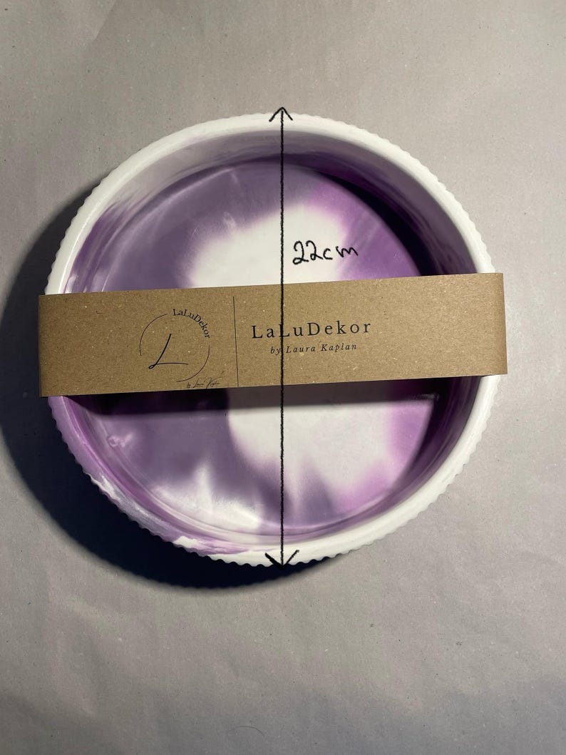 Op de afbeelding: Een ronde decoratieve schaal met een witte, geschulpte rand en een paars en wit gemarmerd interieur. De schaal heeft een diameter van 22 cm. Een bruine papieren band met de tekst "LaLuDekor by Laura Kaplan" is om de schaal gewikkeld.