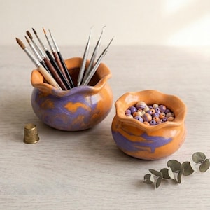Könnte beinhalten: Zwei kleine, handgefertigte Keramikschalen mit gewelltem Rand, bemalt in Orange und Lila. Eine Schale enthält Pinsel und Werkzeuge, die andere ist mit Perlen gefüllt. Ein goldener Fingerhut und ein Zweig Grünzeug sind ebenfalls vorhanden.