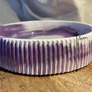 Op de afbeelding: Een ronde, decoratieve kom met een geribbelde buitenkant en een gemarmerd paars en wit interieur. De kom is ongeveer 5 cm hoog. De rand is wit en de kom staat op een houten oppervlak.
