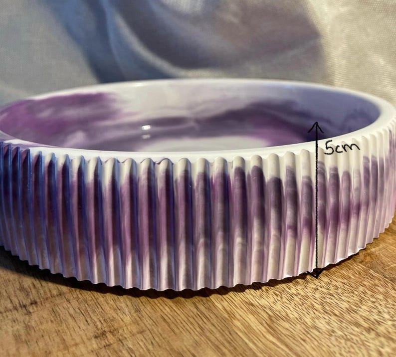 Op de afbeelding: Een ronde, decoratieve kom met een geribbelde buitenkant en een gemarmerd paars en wit interieur. De kom is ongeveer 5 cm hoog. De rand is wit en de kom staat op een houten oppervlak.