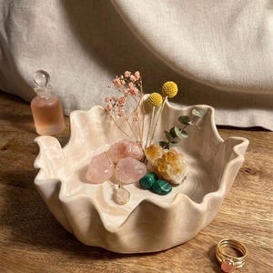 Könnte beinhalten: Eine cremefarbene, gewellte Keramikschale gefüllt mit rosa und gelben Kristallen, getrockneten Blumen und einer goldenen Halskette. Eine kleine Glasflasche und goldene Ringe mit rosa Steinen sind ebenfalls vorhanden. Die Schale steht auf einer Holzoberfläche.