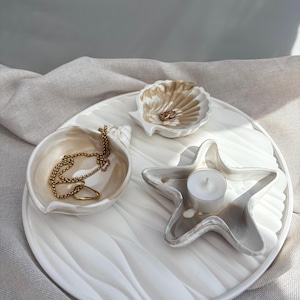 Könnte beinhalten: Ein weißes Keramiktablett mit Wellenmuster hält drei dekorative Muschelschalen. Eine Muschelschale enthält eine goldene Halskette und einen Ring, eine andere zwei Ringe und die dritte eine kleine weiße Kerze. Das Tablett steht auf einem neutralfarbenen Stoff.