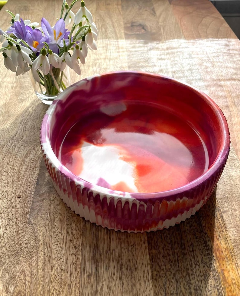 Op de afbeelding: Een ronde, decoratieve kom met een levendig, rood, roze en wit gemarmerd interieur. De kom heeft een getextureerde, geribbelde buitenkant met een wit en roze patroon. Een kleine glazen vaas met paarse en witte bloemen staat in de buurt.