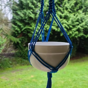 Puede incluir: Un macetero de macramé azul con una maceta de cerámica blanca. El macetero está colgado de un gancho de techo y la maceta está vacía.