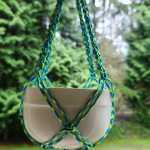 Puede incluir: Una maceta de cerámica blanca colgando de un macramé para plantas con cordones azules y amarillos. El macramé está suspendido de un gancho de techo.