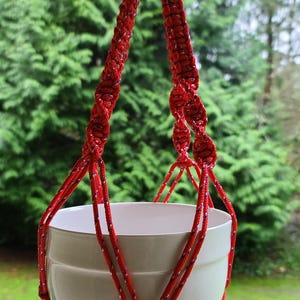 May include: Red macrame plant hanger with a white ceramic pot. The hanger is made of thick rope and has a tassel at the bottom.