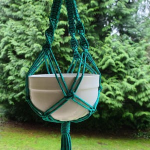 May include: A teal macrame plant hanger with a white ceramic pot hanging from a metal hook. The hanger is made of thick rope and has a tassel at the bottom.