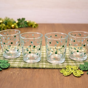 May include: Four clear glass votive candle holders with green and blue polka dots are arranged on a green and white checkered placemat. Decorative shamrocks and a green garland are also present, creating a festive St. Patrick's Day theme.