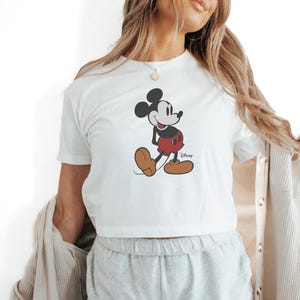 May include: White t-shirt featuring a classic Mickey Mouse graphic. The cartoon character is depicted in black, red, and brown. The Disney logo is printed in black on the bottom right. The shirt is paired with gray shorts.