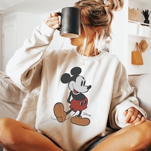 Pu&ograve; includere: Una felpa beige con una grafica di Topolino. Il personaggio dei cartoni animati &egrave; raffigurato nella sua posa iconica, con orecchie nere, viso bianco, pantaloncini rossi e scarpe marroni. Il logo Disney &egrave; stampato sotto.