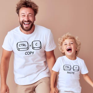 以下が含まれることがあります： 黒いグラフィックが入ったお揃いの白いTシャツを着た2人。1つのシャツには「CTRL + C COPY」、もう1つには「CTRL + V PASTE」と書かれています。シャツは半袖です。