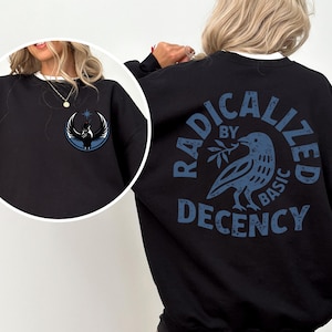 May include: Black sweatshirt with a white collar. The front has a circular emblem with a bird design. The back displays the text "RADICALIZED BY BASIC DECENCY" around a blue bird holding a branch. A casual, comfortable garment.