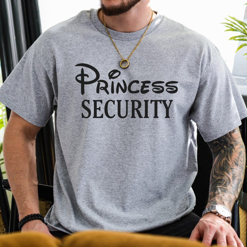 Puede incluir: Una camiseta gris con un gr&aacute;fico negro que dice "Princess Security" en una fuente estilizada. El gr&aacute;fico recuerda a un logotipo de Disney.