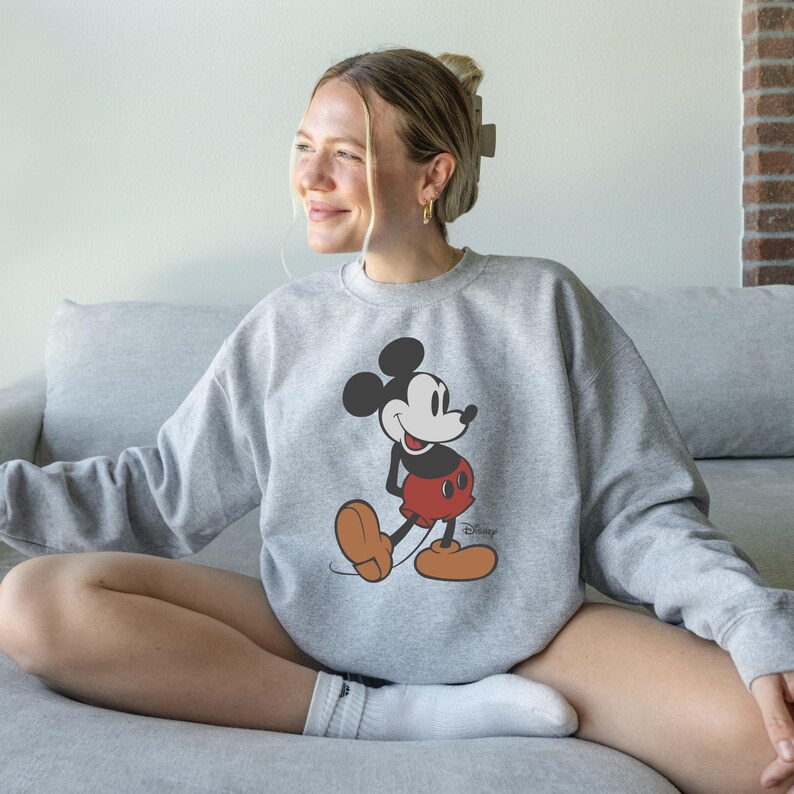 Pu&ograve; includere: Una felpa grigia con un disegno di Topolino. Il personaggio dei cartoni animati &egrave; raffigurato nella sua posa classica, con pantaloncini rossi, scarpe marroni e guanti bianchi. Il logo Disney &egrave; visibile in basso a destra.