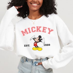 May include: White sweatshirt featuring the word "MICKEY" in pink block letters above a cartoon image of Mickey Mouse. The sweatshirt also includes the text "EST. 1928" in black. The design is centered on the front of the long-sleeved top.