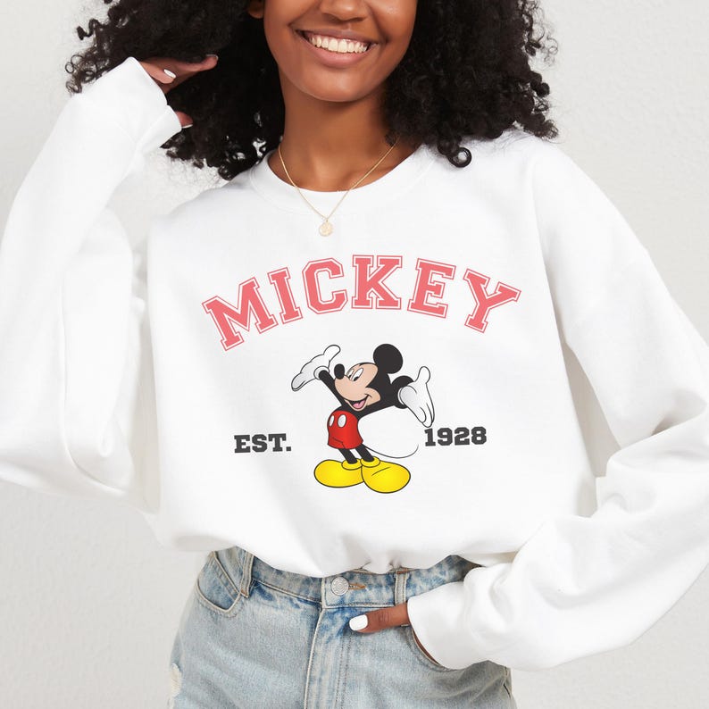 May include: White sweatshirt featuring the word "MICKEY" in pink block letters above a cartoon image of Mickey Mouse. The sweatshirt also includes the text "EST. 1928" in black. The design is centered on the front of the long-sleeved top.